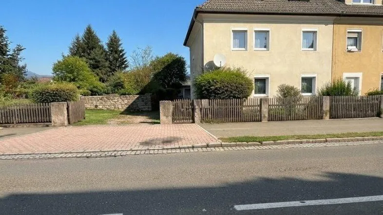 Ansicht des Hauses von der Straße mit Vorgarten und Zaun unter blauem Himmel.