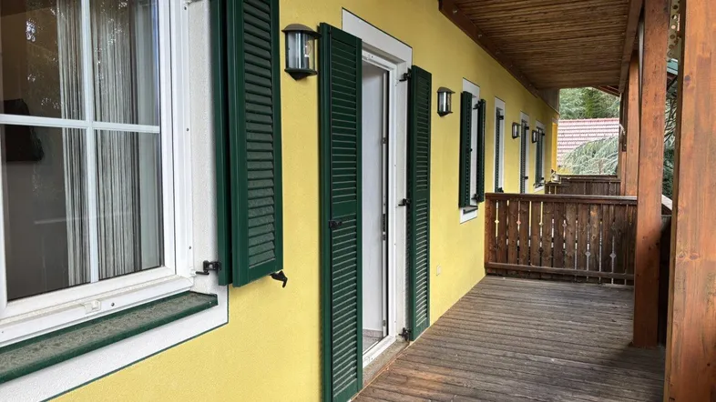 Überdachter Balkon mit Holzboden und grünen Fensterläden, bietet Zugang zu den Innenräumen.
