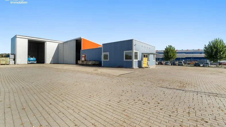 Blick auf das weitläufige Außengelände mit mehreren Industriehallen und einem Bürogebäude unter blauem Himmel.