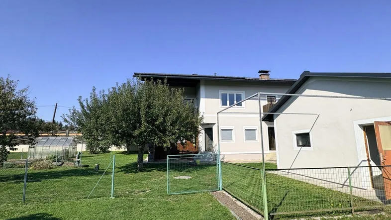 Hausfassade mit Garten, Obstbäumen und einem Nebengebäude unter blauem Himmel.