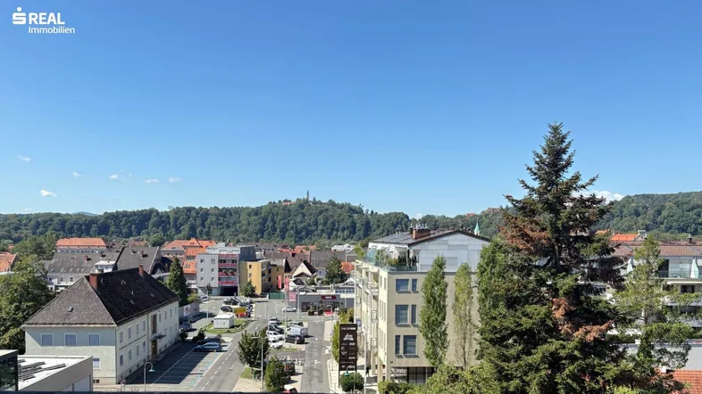 Weitläufiger Blick über eine Stadt mit Gebäuden, einer Straße und einem bewaldeten Hügel unter klarem blauem Himmel.