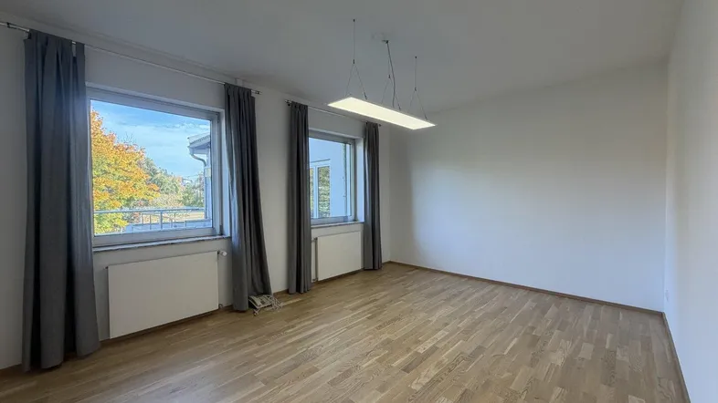 Heller Wohnraum mit Holzboden, weißen Wänden und zwei Fenstern, die viel Tageslicht hereinlassen.