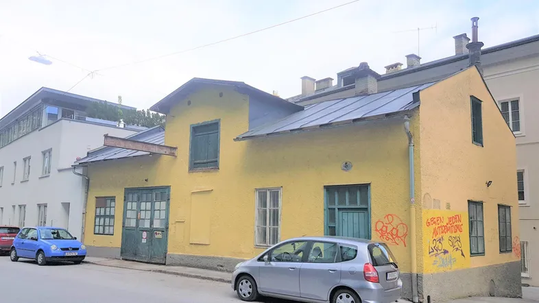 Gelbes Gebäude mit grünen Türen und Fenstern an einer Straße, mit parkenden Autos davor.