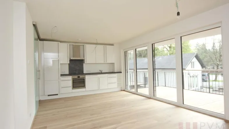 Heller offener Wohnbereich mit integrierter weißer Küche, Holzboden und Zugang zu einem großen Balkon.