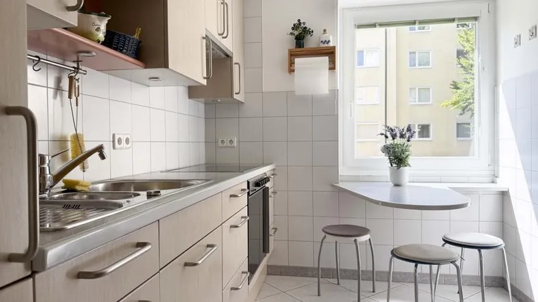 Funktionale Küche mit hellen Holzfronten, weißem Fliesenboden und kleinem Essbereich am Fenster.
