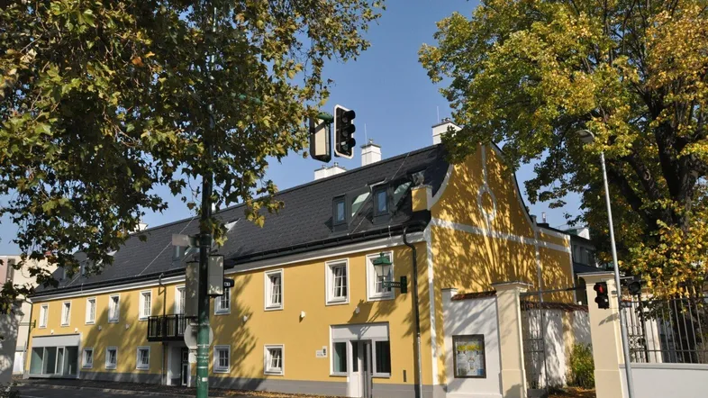 Gelbes Gebäude mit traditioneller Architektur, vielen Fenstern und umgebenden Bäumen an einem sonnigen Tag.
