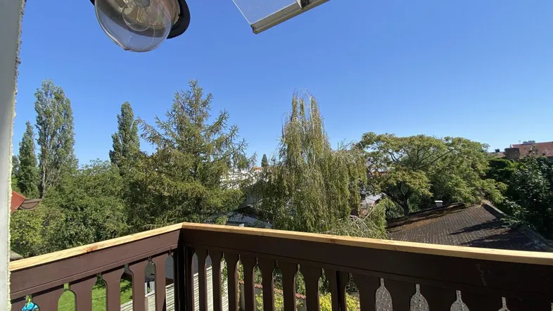 Balkon mit Holzbalkongeländer und Außenleuchte, bietet Blick auf Bäume und Dächer.