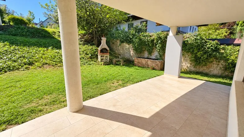 Überdachte Terrasse mit Fliesenboden und gemauertem Grill im grünen Gartenbereich.