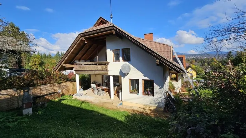 Charmantes Einfamilienhaus mit überdachter Terrasse, Garten und Blick auf die umliegende Landschaft unter blauem Himmel.