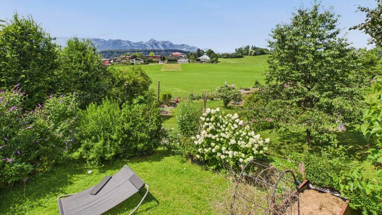Grüner Garten mit Liegestuhl, blühenden Büschen und weitem Blick auf die Landschaft und Berge.