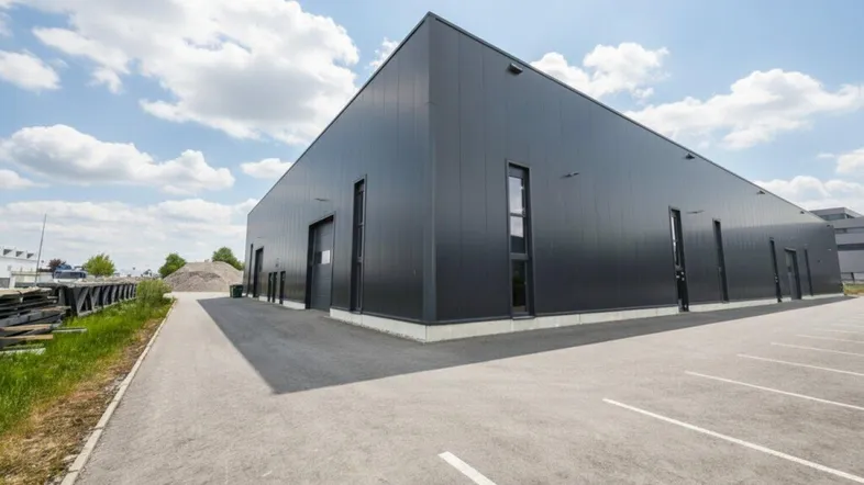 Moderne Gewerbehalle mit dunkler Fassade und großem Parkplatz unter blauem Himmel.