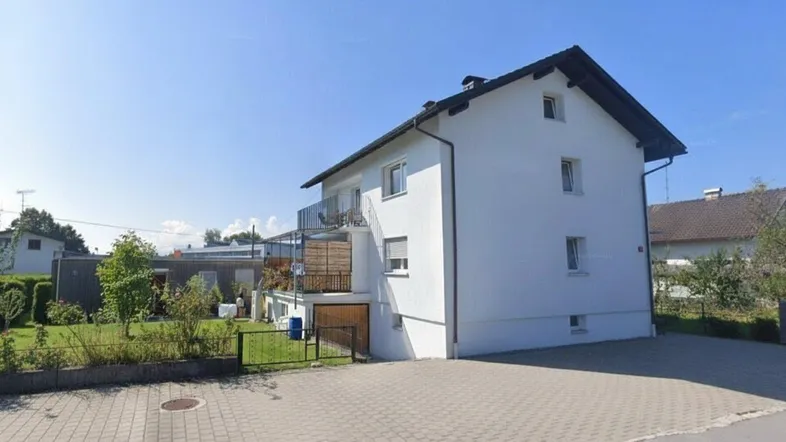 Weißes Einfamilienhaus mit Satteldach, Balkonen und gepflasterter Einfahrt unter blauem Himmel.