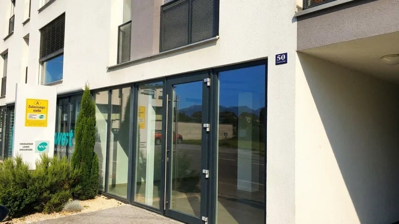 Moderne Gebäudefassade mit großen Glasfenstern und Eingangstüren, umgeben von Grünflächen.