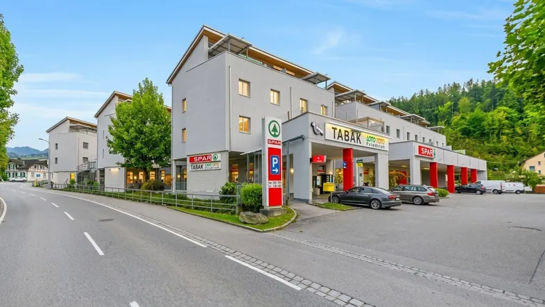 Modernes Gebäude mit Geschäften im Erdgeschoss und Wohnungen darüber, umgeben von grüner Landschaft.