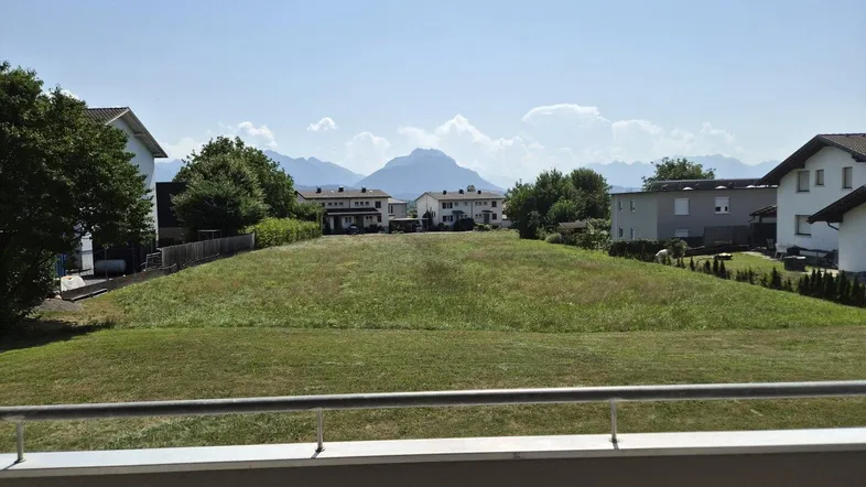 Blick vom Balkon auf eine grüne Wiese mit umliegenden Häusern und einer beeindruckenden Bergkulisse im Hintergrund.