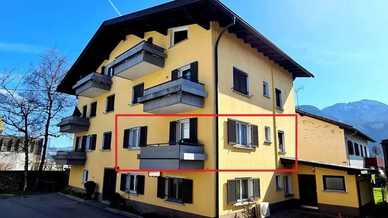 Gelbes Mehrfamilienhaus mit mehreren Balkonen und dunklen Fensterläden vor einer Bergkulisse.