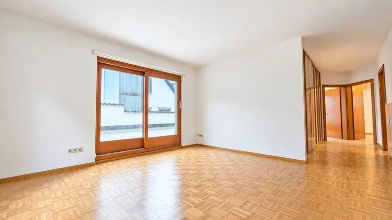 Helles, leeres Zimmer mit Parkettboden und Zugang zu einem Außenbereich durch eine große Schiebetür.