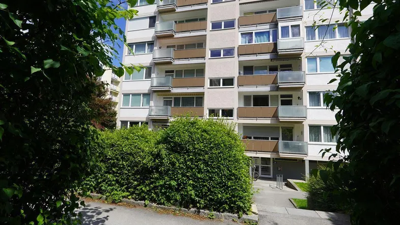 Mehrstöckiges Wohngebäude mit Balkonen, umgeben von grünen Hecken und Bäumen.