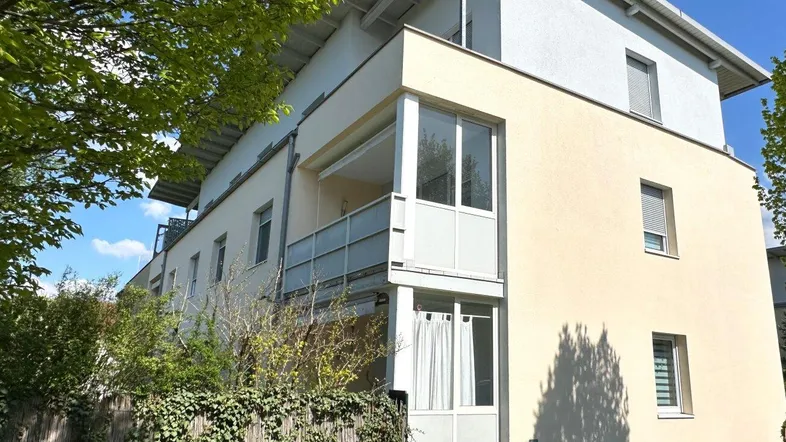 Mehrfamilienhaus mit heller Fassade, Balkonen und umgebender Grünfläche unter blauem Himmel.