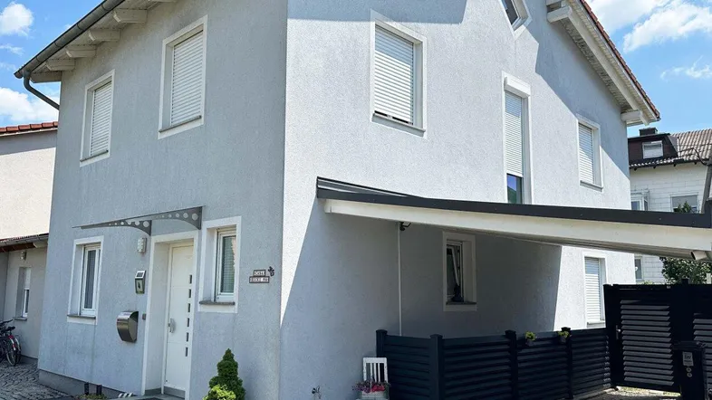 Graues Einfamilienhaus mit Rollläden und überdachtem Eingangsbereich unter blauem Himmel.
