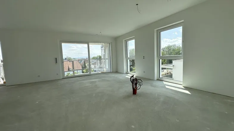 Helles, unfertiges Wohnzimmer mit großen Fenstern, Betonboden und Blick auf die Umgebung.