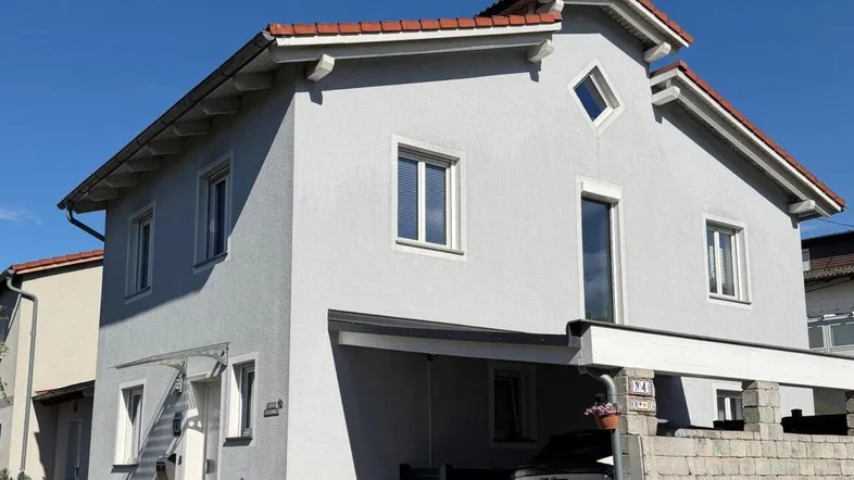 Modernes Einfamilienhaus mit hellgrauer Fassade, rotem Ziegeldach und Carport an einem sonnigen Tag.