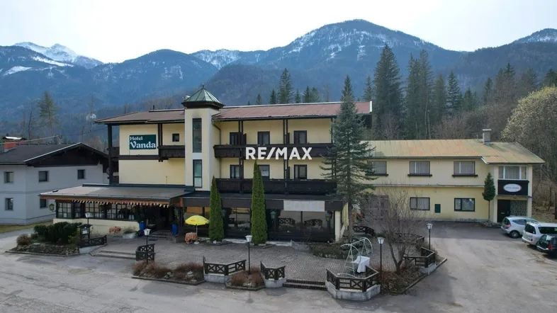 Traditionelles Hotelgebäude mit Parkplatz vor einer beeindruckenden Bergkulisse und grüner Umgebung.