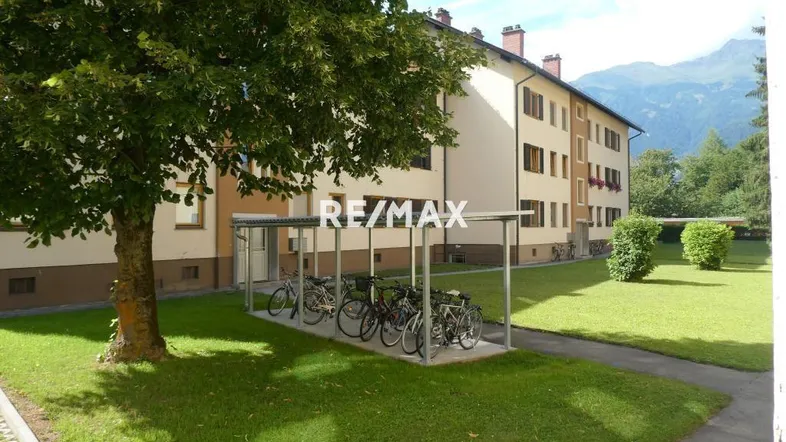 Mehrfamilienhaus mit Fahrradunterstand und gepflegter Grünfläche vor alpiner Kulisse.