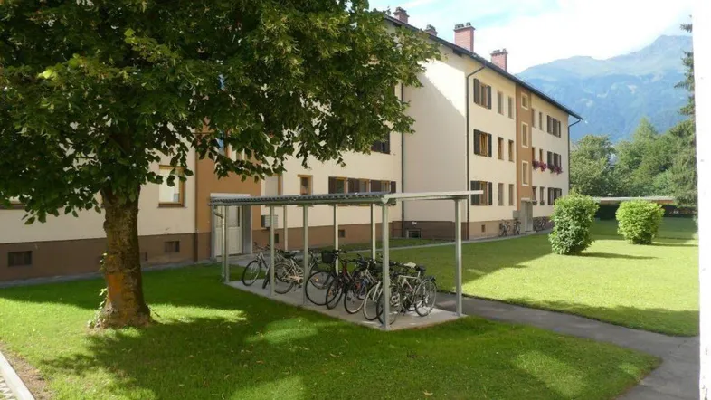 Mehrfamilienhaus mit Fahrradunterstand und gepflegter Grünfläche, umgeben von Bergen.