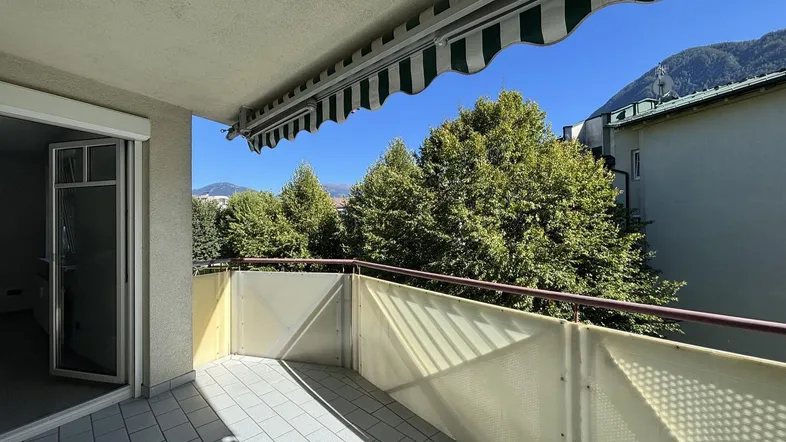 Heller Balkon mit Fliesenboden, Markise und Blick auf grüne Bäume und Berge.