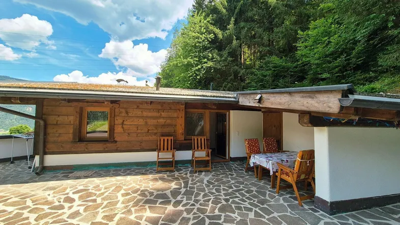 Große gepflasterte Terrasse mit Holzhütte und Sitzgelegenheiten, umgeben von grünen Bäumen und Bergen.