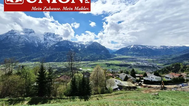 Panoramablick auf schneebedeckte Berge und eine grüne Landschaft mit Wohnbebauung unter blauem Himmel.