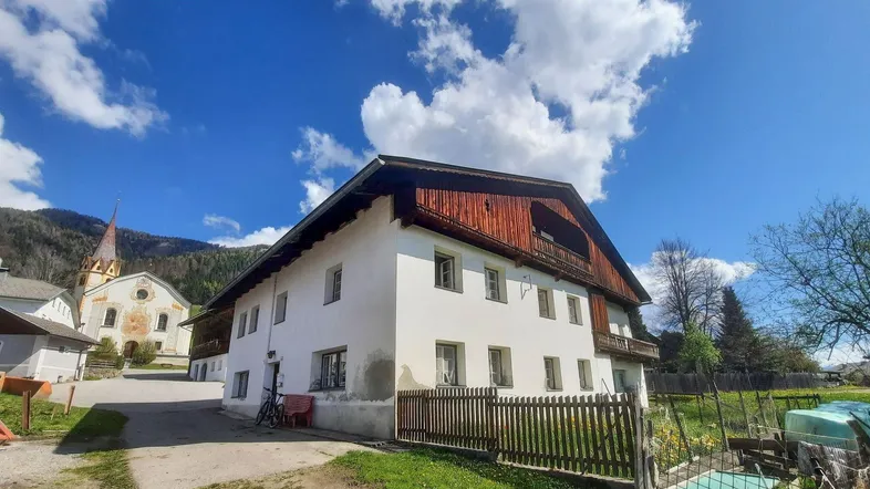 Traditionelles Haus mit weißer Fassade und Holzbalkon, umgeben von ländlicher Landschaft und einem Kirchturm im Hintergrund.