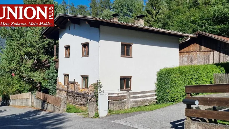 Weißes Wohnhaus mit Holzelementen und grüner Hecke in ländlicher Umgebung.