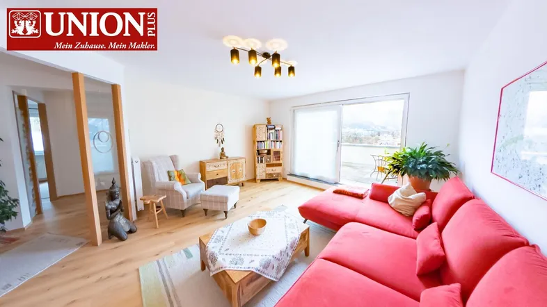 Helles Wohnzimmer mit rotem Sofa, Parkettboden und Zugang zu einem Balkon mit Bergblick.