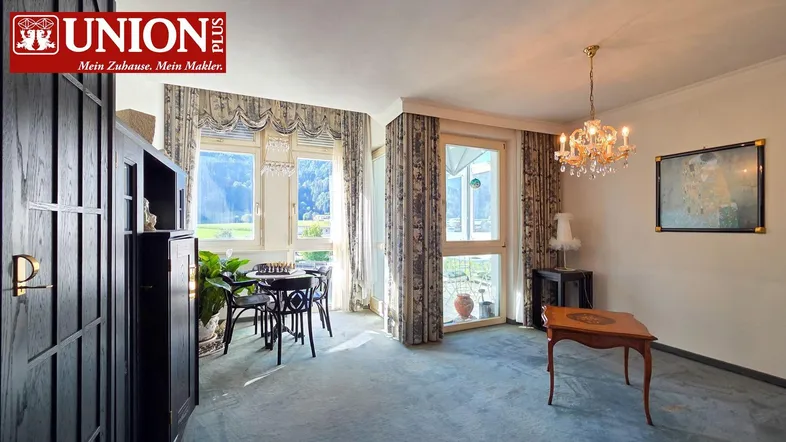 Heller Wohn- und Essbereich mit Teppichboden, Kronleuchter und Bergblick durch große Fenster.