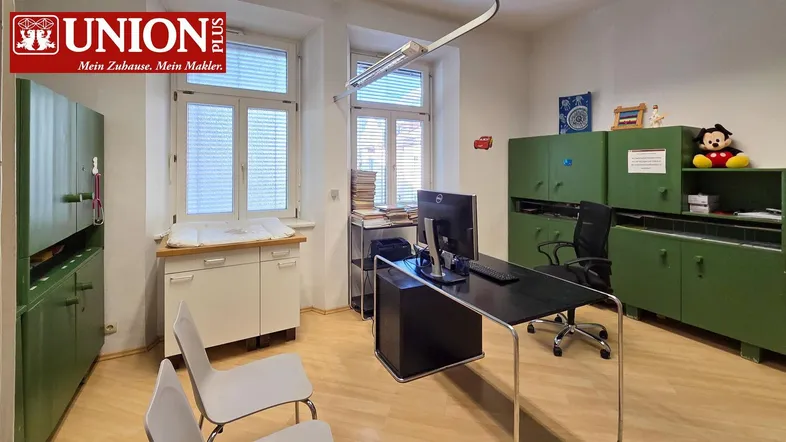 Heller Raum mit Schreibtisch, Computer und grünen Schränken, ideal als Büro oder Behandlungszimmer.