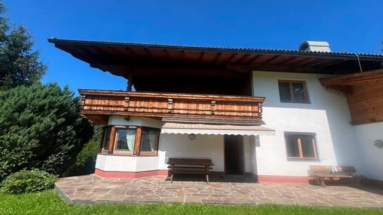 Traditionelles Haus mit großer gepflasterter Terrasse und Holzbalkon, umgeben von Grünflächen.
