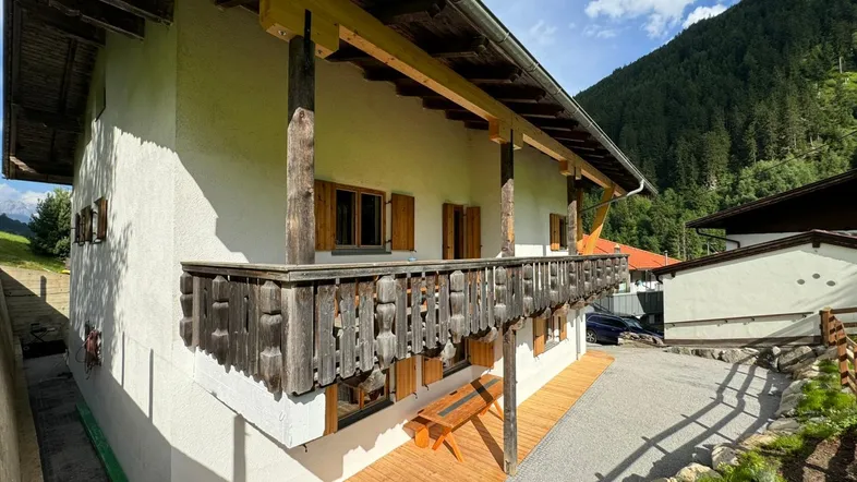 Traditionelles Haus im alpinen Stil mit Holzbalkon und Terrasse, umgeben von grünen Bergen.