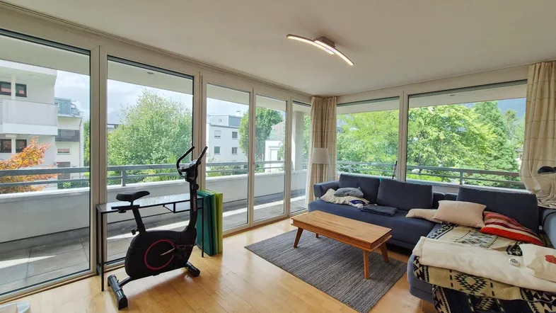 Helles Wohnzimmer mit Holzboden, bequemer Couch und Zugang zu einem breiten Balkon mit grüner Aussicht.