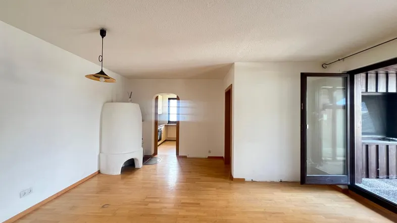 Helles Wohnzimmer mit Holzboden, traditionellem Kachelofen und Zugang zu einem Außenbereich.