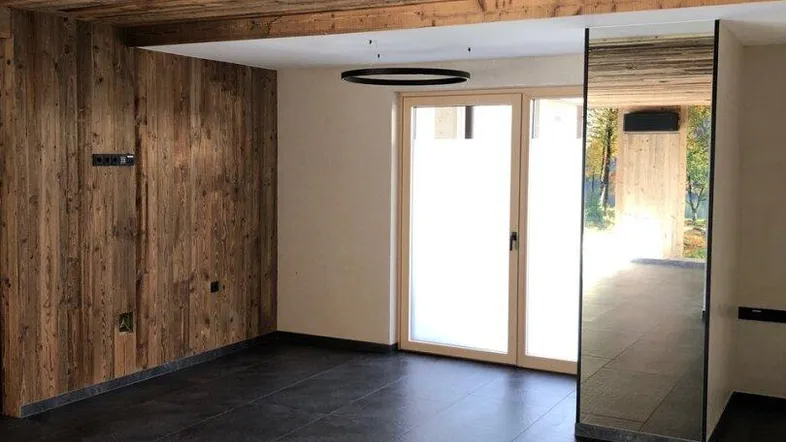 Geräumiger Wohnbereich mit Holzwänden und -decke, dunklen Fliesen und großen Fenstern, die viel Licht hereinlassen.