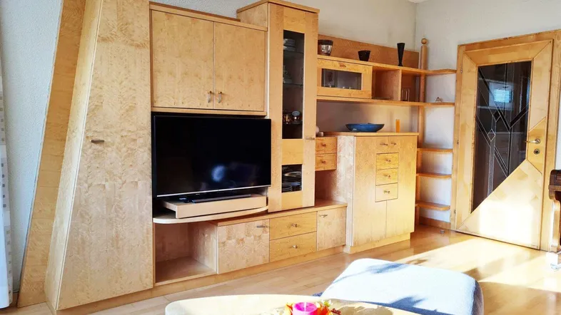 Gemütliches Wohnzimmer mit heller Holzschrankwand, Fernseher und einem bequemen Sessel.