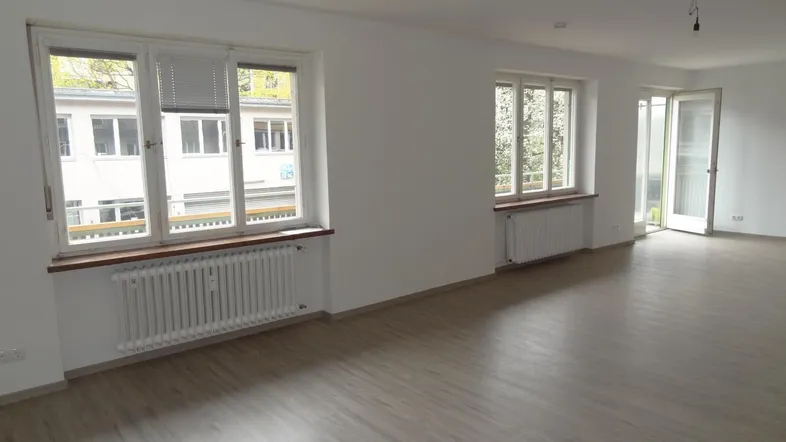 Heller, leerer Raum mit großen Fenstern, die viel Tageslicht hereinlassen, und modernem Laminatboden.