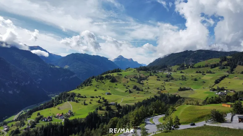 Panoramablick auf die grüne Berglandschaft mit verstreuten Häusern und kurvigen Straßen unter blauem Himmel.