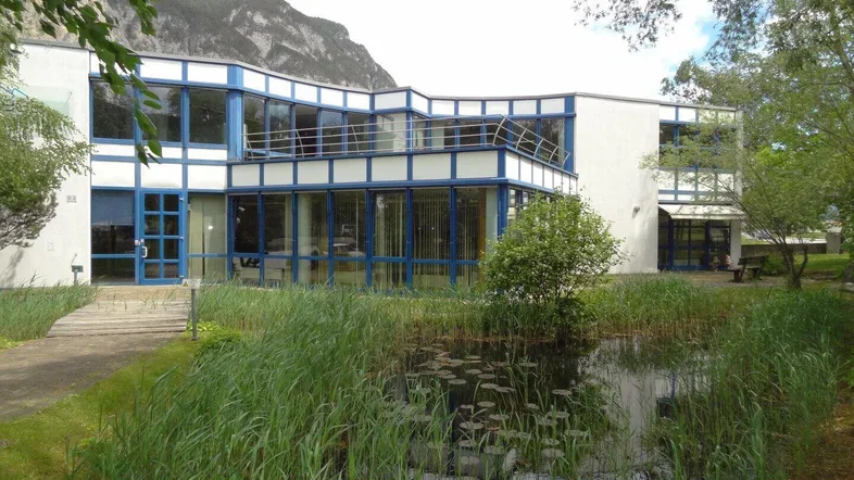 Modernes Bürogebäude mit blauen Fensterrahmen, umgeben von Grünflächen und einem Teich mit Bergblick.