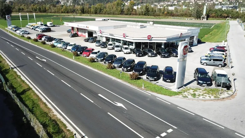Luftaufnahme eines Autohauses mit großem Parkplatz, umgeben von grünen Feldern und Bergen im Hintergrund.