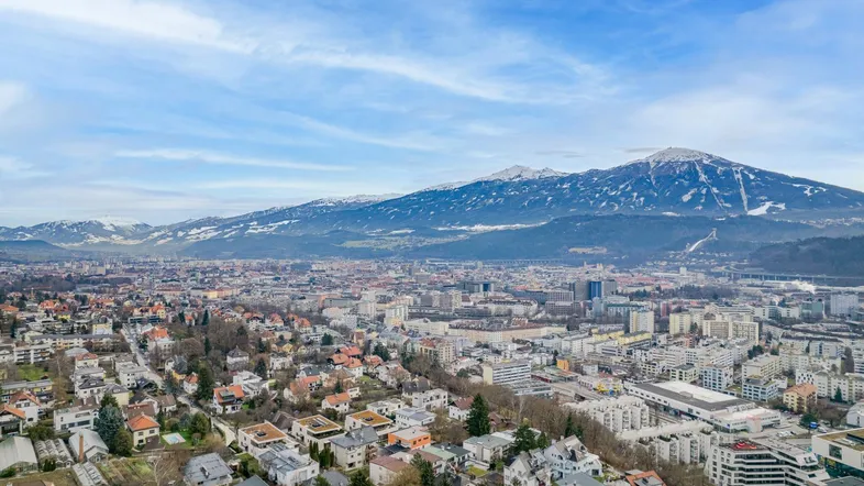Weitläufige Luftaufnahme einer Stadtlandschaft mit schneebedeckten Bergen im Hintergrund unter blauem Himmel.