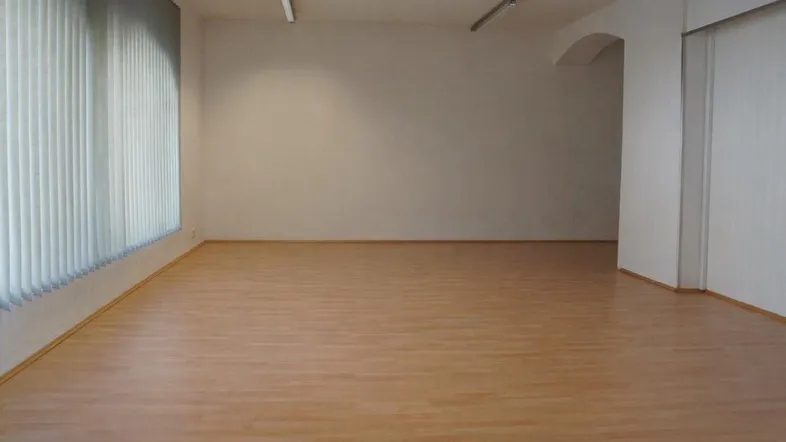 Leerer Raum mit hellem Laminatboden, weißen Wänden und vertikalen Jalousien am großen Fenster.
