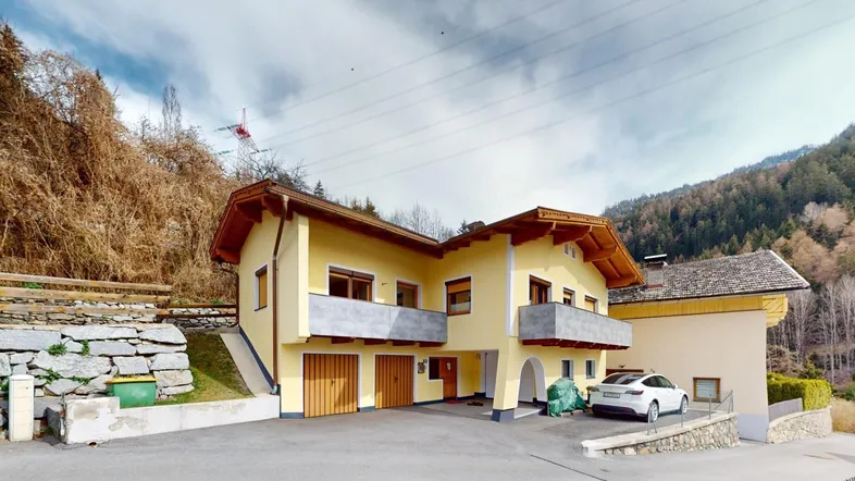 Gelbes Wohnhaus mit Balkonen, Garagen und einem Auto auf der Einfahrt vor bergiger Kulisse.